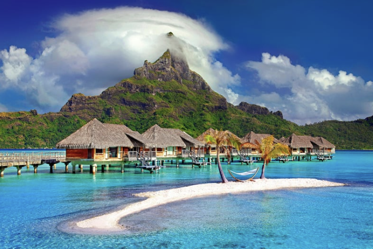 Overwater bungalows with thatched roofs above turquoise Caribbean waters, a hammock between palm trees on a small white-sand islet, and a lush green mountain with clouds in the background.